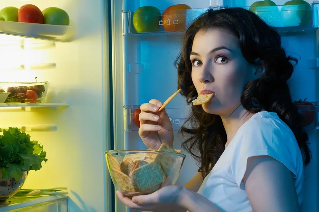Woman looking surprised while eating crackers in an open fridge.